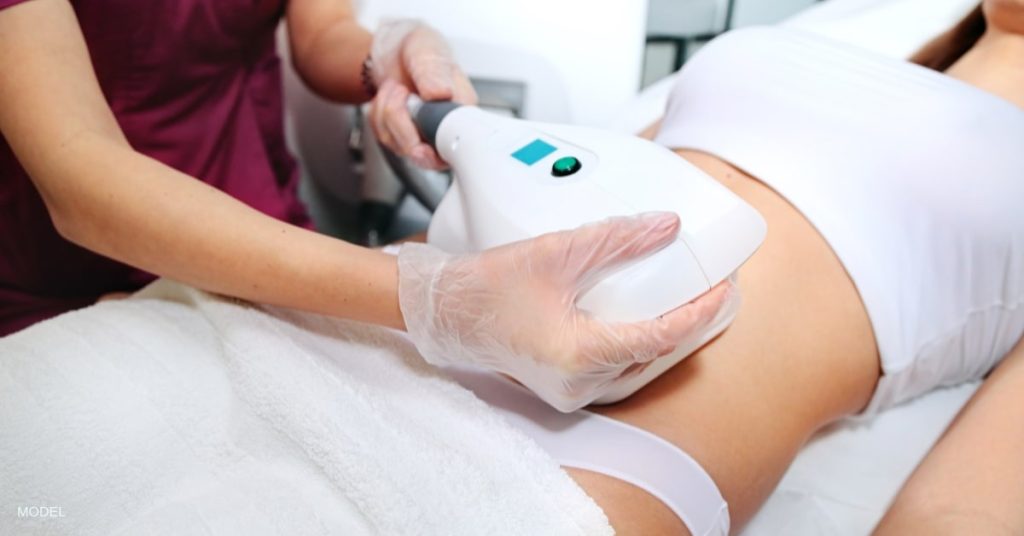 In a treatment room, a woman is receiving a CoolScultping® treatment on her abdomen from a nurse who is holding the device. (models)