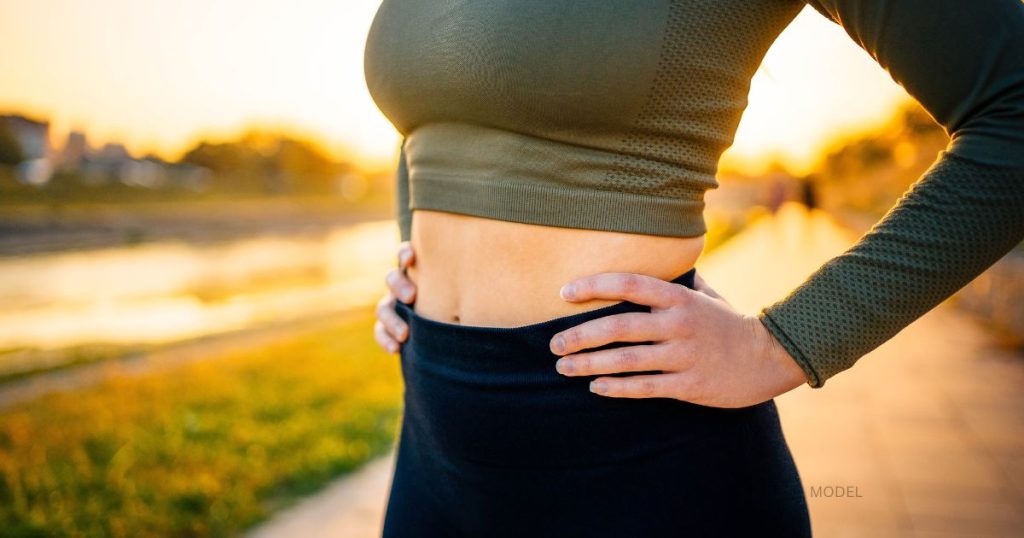 Woman (model) with toned abs and hands on hips outdoors during sunset.